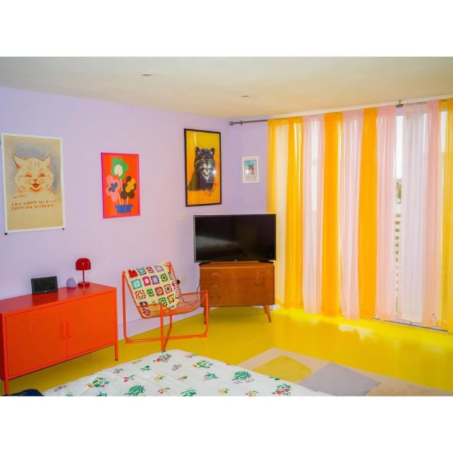 Retro-style interior with lilac walls, yellow floor, orange furniture, colorful posters, and sheer curtains in a brightly lit room.