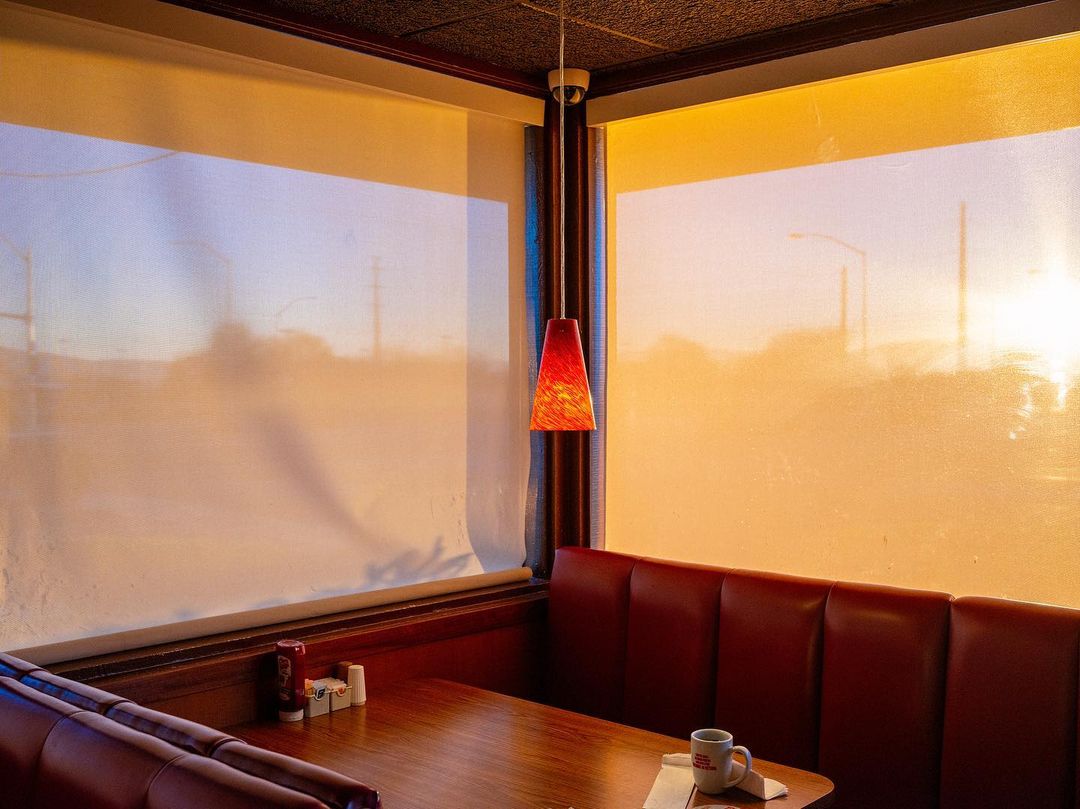 Diner booth seating with red benches, wood table, a mug, and sunlight shining through the large windows casting a warm golden glow.