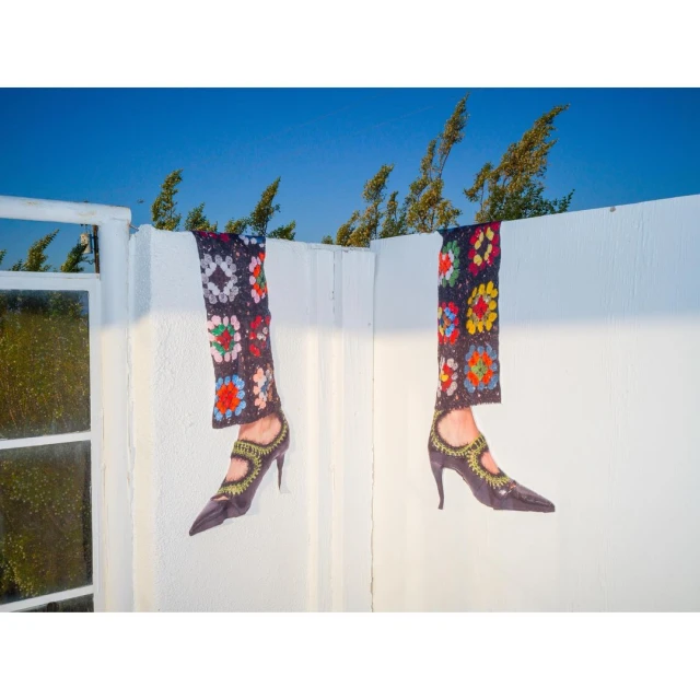 A surreal outdoor scene featuring two pairs of disembodied legs adorned in colorful crocheted textiles and high-heeled shoes against a blue sky.