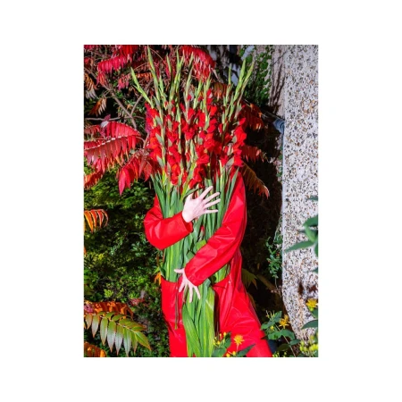 Person in a red jumpsuit stands in a garden hugging a large bouquet of red gladiolus flowers with lush green foliage in daytime lighting.