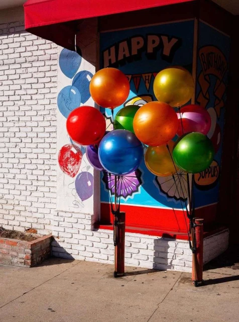 A cluster of colorful balloons is tethered outside a brick building with a red awning and whimsical balloon mural for a celebration.