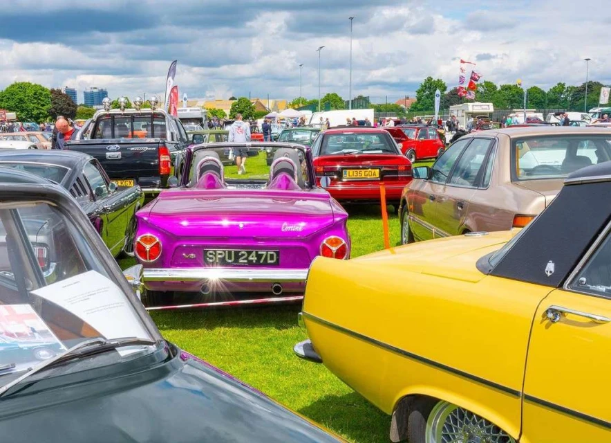 A collection of classic cars in various colors, including purple, yellow, and red, are displayed on a green lawn under a cloudy sky.