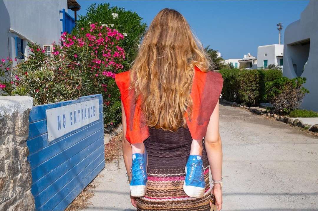 Rear view of an adult woman carrying a child in a carrier through a white-washed village, with blooming flowers and blue accents.
