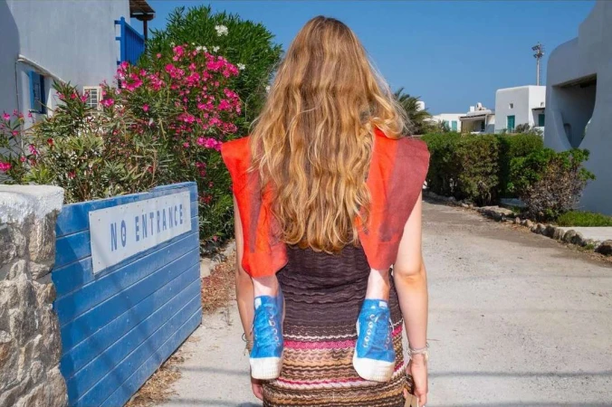 Rear view of an adult woman carrying a child in a carrier through a white-washed village, with blooming flowers and blue accents.