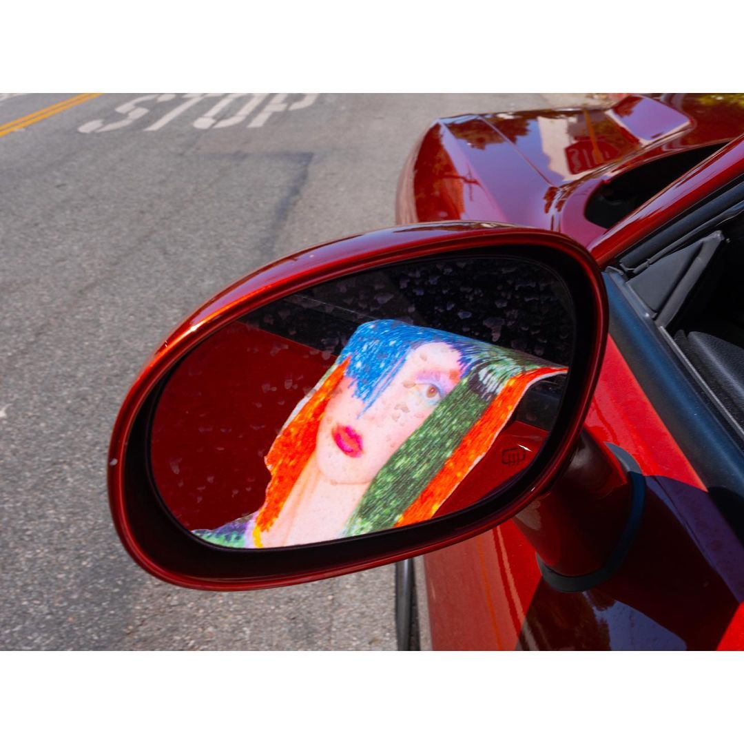 Close-up of red sports car side mirror reflecting a colorful art drawing of a person with blue hair, red lips, and orange scarf.