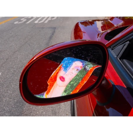 Close-up of red sports car side mirror reflecting a colorful art drawing of a person with blue hair, red lips, and orange scarf.