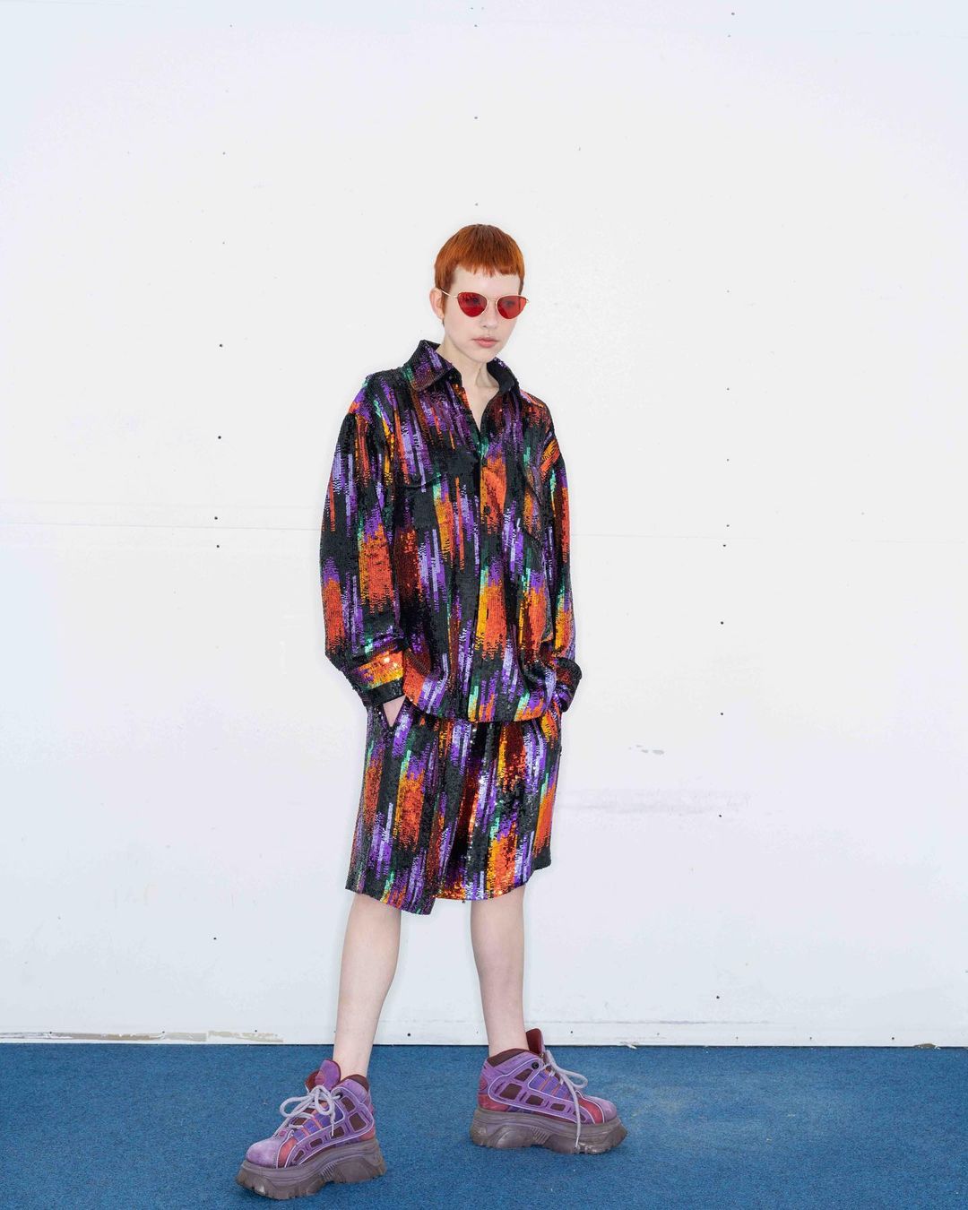 A person stands against a white wall wearing a matching black, orange, and purple brush-stroke patterned shirt and shorts, with red sunglasses.