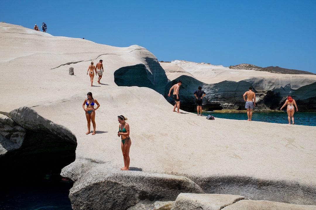 People relax on white rock formations by the sea under a clear sky. Some sunbathe, others stand or walk near the water's edge.