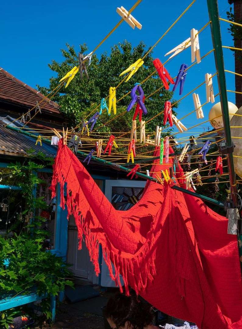 Outdoor laundry scene with colorful clothespins on lines, red fabric hanging, green foliage, blue sky, and a rustic architectural backdrop.