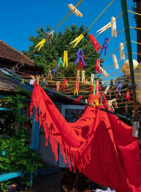 Outdoor laundry scene with colorful clothespins on lines, red fabric hanging, green foliage, blue sky, and a rustic architectural backdrop.