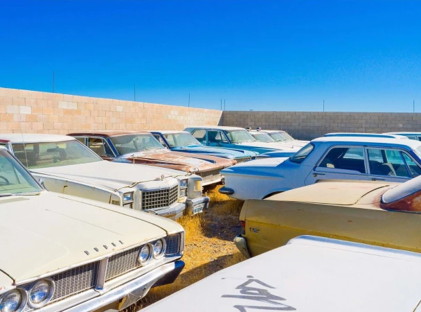 Several aged, abandoned cars are densely packed in a junkyard under a clear blue sky, with a tan brick wall marking the background.