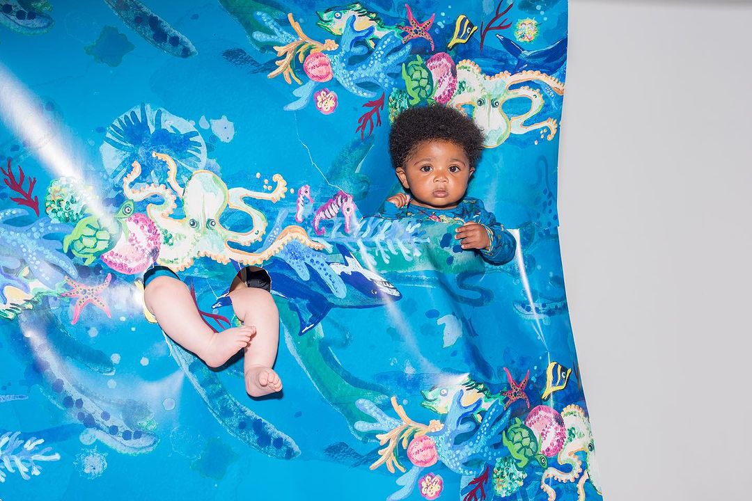 Infant with Afro poses behind a blue ocean-themed graphic with octopus, dolphins, sea horses, and starfish in a studio setting.