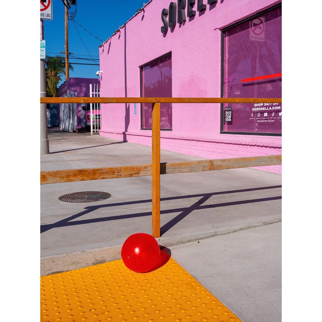 Pink building with storefront windows is shown beside a sidewalk. A red ball rests on the textured yellow safety tile of a sidewalk.