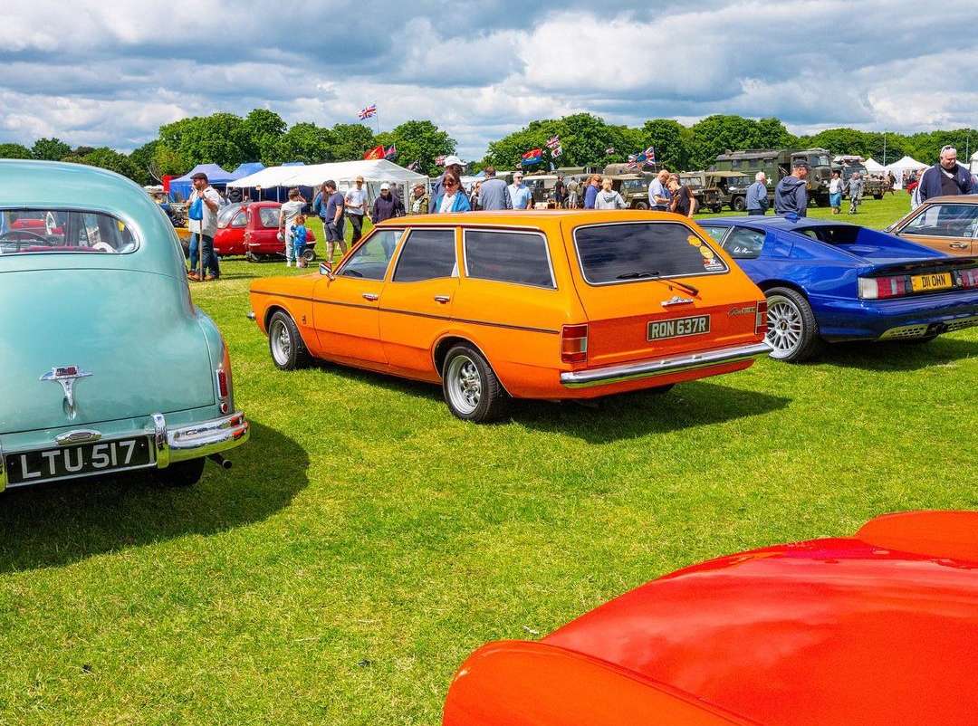 Classic orange station wagon parked on green grass at an outdoor car show, surrounded by people and other vintage automobiles on display.