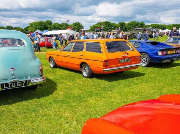 Classic orange station wagon parked on green grass at an outdoor car show, surrounded by people and other vintage automobiles on display.