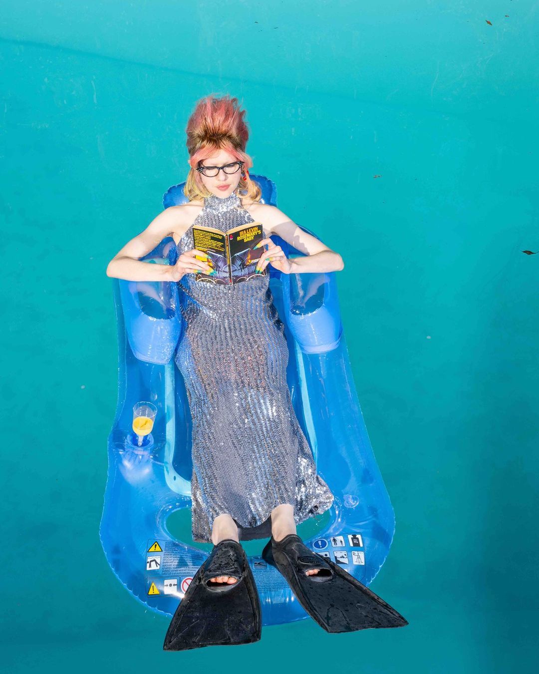 Woman in sparkly dress, flippers, and inflatable lounger reads a book in a blue pool on a sunny day, creating a whimsical, eye-level image.