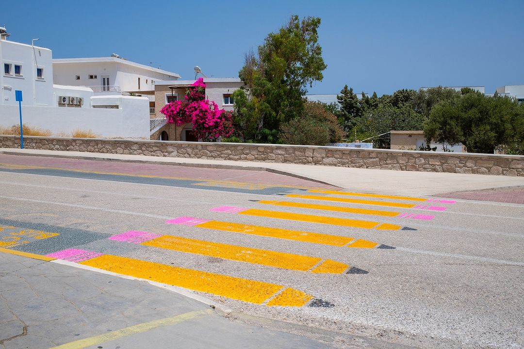 Eye-level shot of a crosswalk with painted pencil design on an asphalt road in a sunny, outdoor setting, with white buildings in the background.