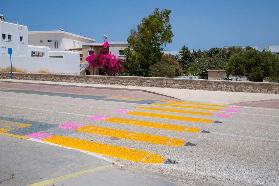 Eye-level shot of a crosswalk with painted pencil design on an asphalt road in a sunny, outdoor setting, with white buildings in the background.