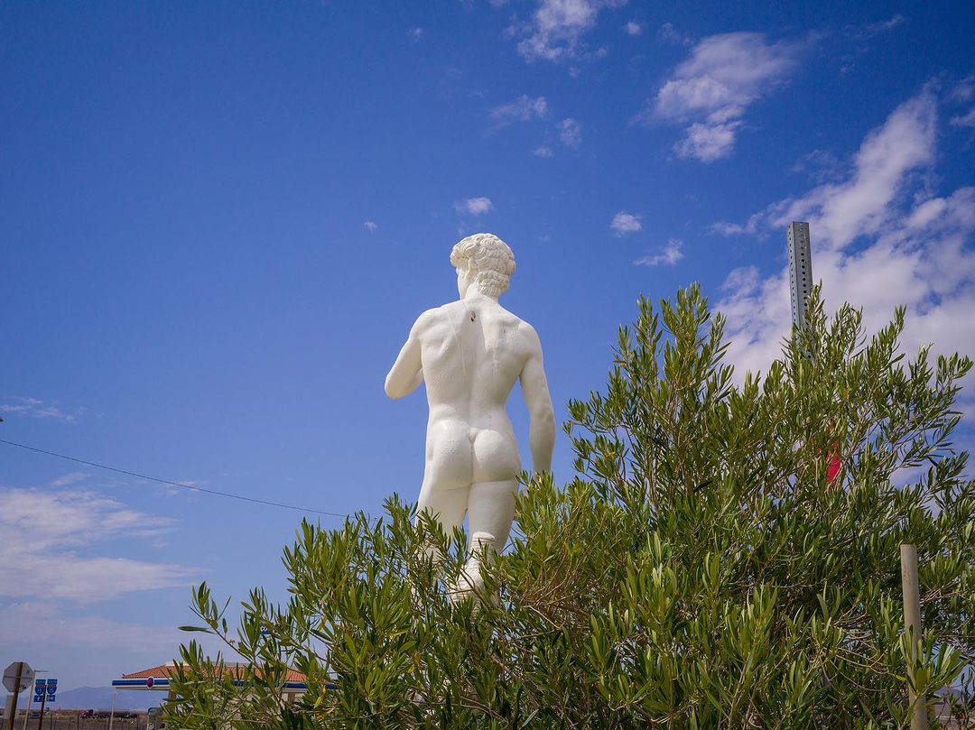 Statue of a nude male figure set against a partly cloudy sky, partially obscured by a leafy green bush in an outdoor setting.