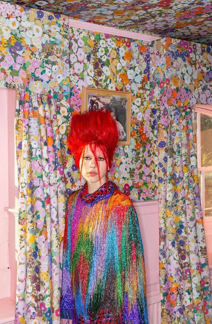 A person with bright red hair and colorful fringed outfit stands in an eclectic interior filled with floral wallpaper and pink accents.