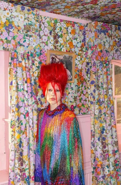 A person with bright red hair and colorful fringed outfit stands in an eclectic interior filled with floral wallpaper and pink accents.