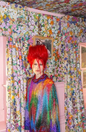 A person with bright red hair and colorful fringed outfit stands in an eclectic interior filled with floral wallpaper and pink accents.