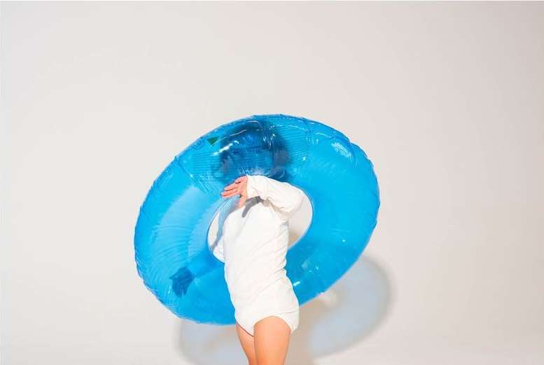 Child in white outfit holds blue inflatable swim ring against a white backdrop in a studio shot with soft, diffused lighting.
