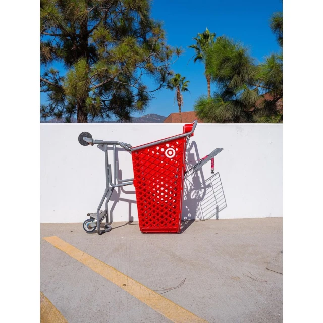 Red shopping cart on its side against a white wall in an outdoor parking lot, set against a backdrop of trees and blue sky.