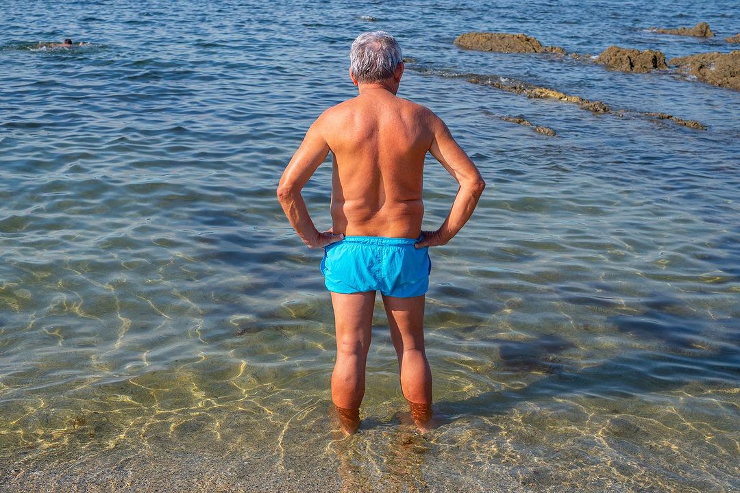 Back view of a senior man in blue swim trunks standing waist-deep in the ocean with rocks in the distance on a sunny day.