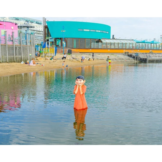 Young child in an orange dress standing in water with hands on face, with beach and buildings in background during daytime.