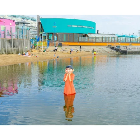 Young child in an orange dress standing in water with hands on face, with beach and buildings in background during daytime.