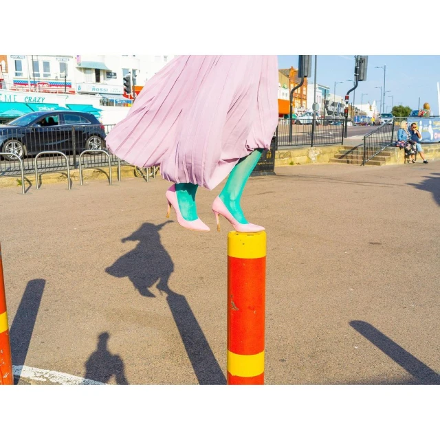 Woman in pink heels and green tights balances on a red and yellow bollard under bright sunlight, casting a long shadow on the asphalt.
