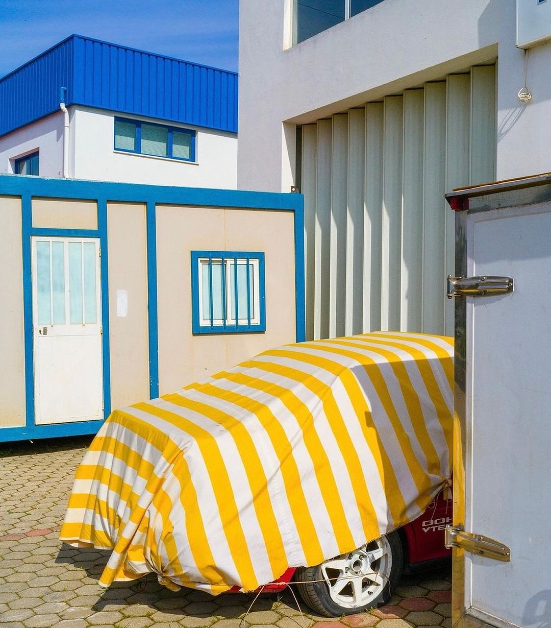 A car is parked in front of a building and covered with a yellow and white striped tarp, partially visible, on a sunny day.