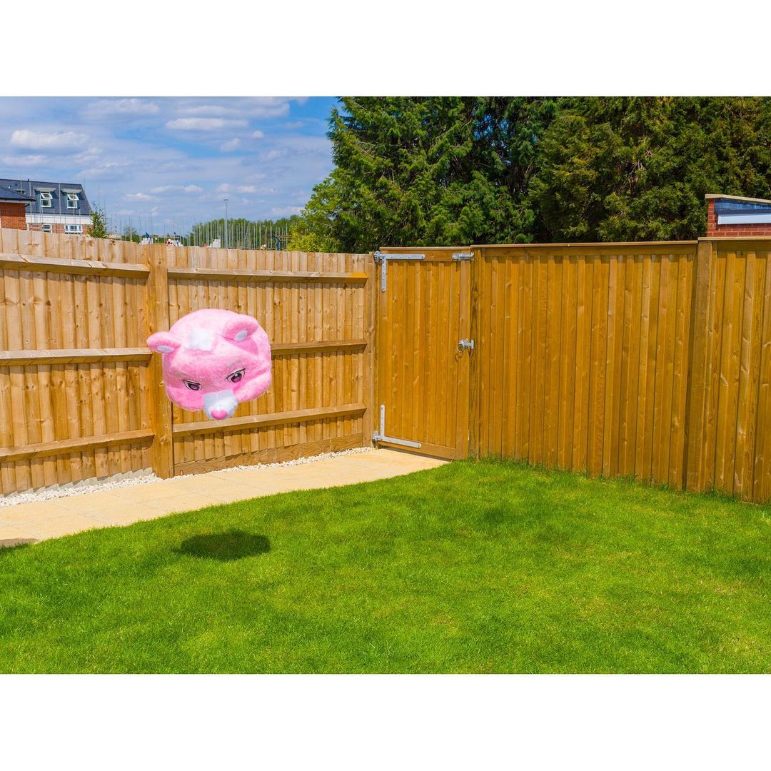 Pink pig-shaped balloon floats in a backyard with a wooden fence and green lawn under a blue sky with white clouds. 
