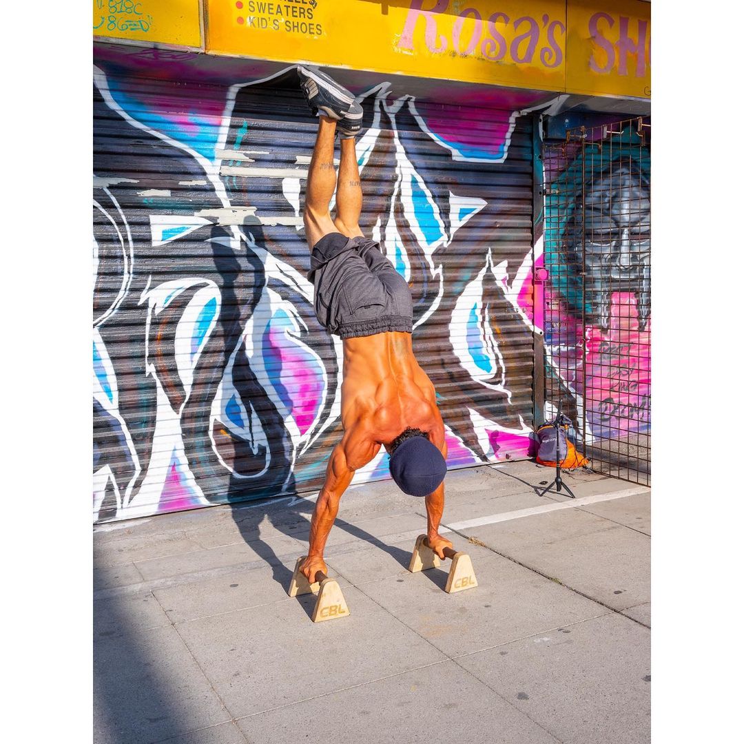 A muscular man in shorts and a baseball cap balances in a handstand on wooden parallettes outdoors against a graffiti wall.