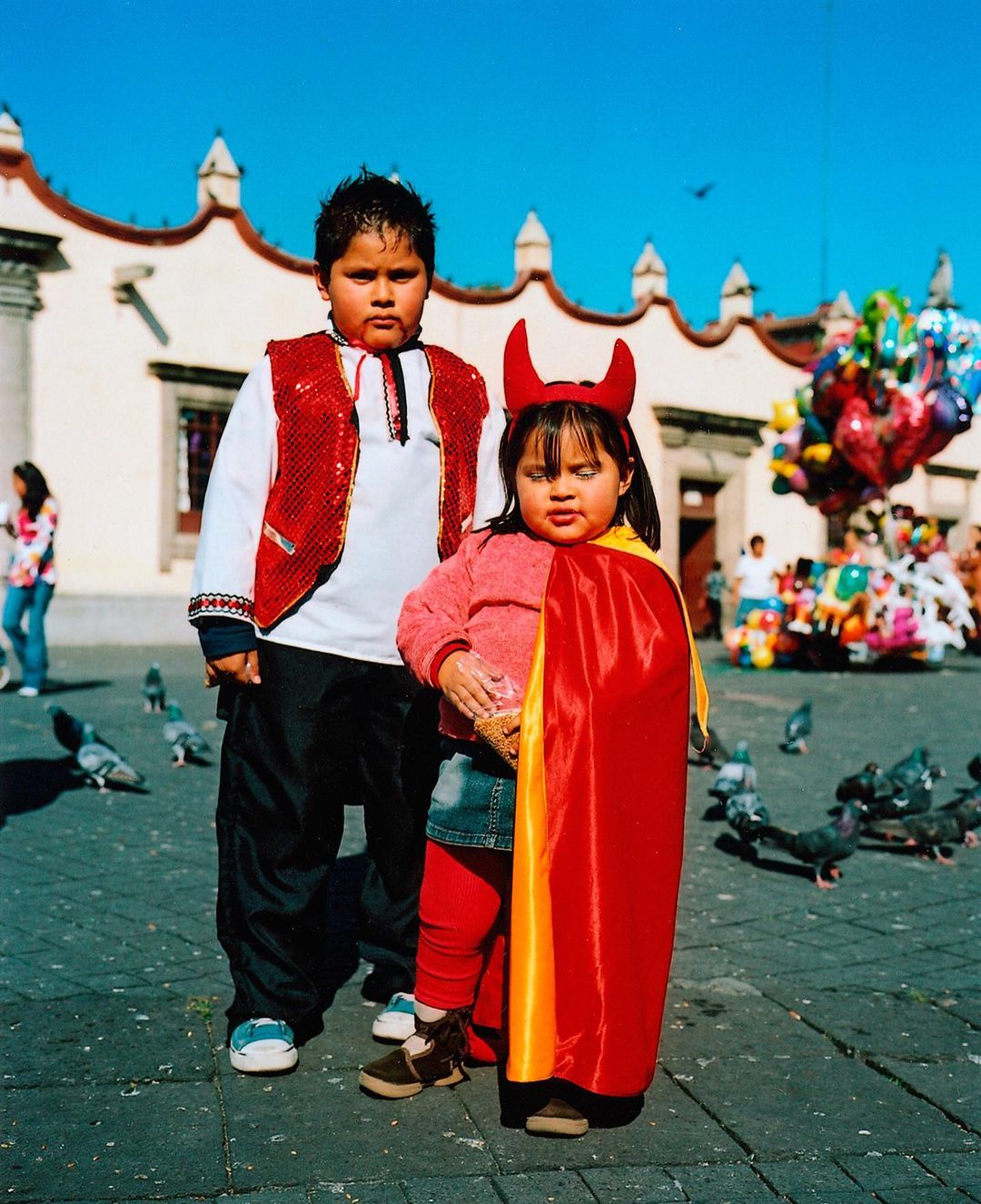 Two children in costume stand in a sunny urban plaza near a building and colorful balloons, with pigeons scattered on the patterned ground.