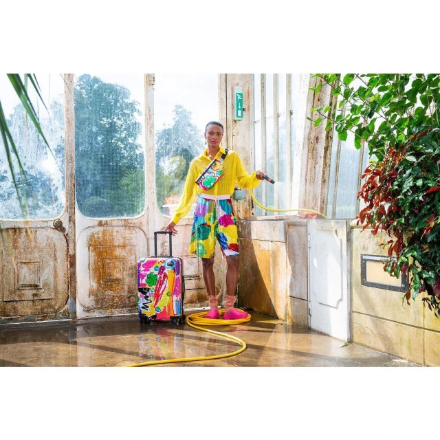 Woman in yellow shirt and colorful shorts stands with a patterned suitcase and yellow hose in a sunlit greenhouse. 