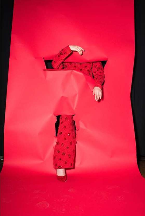 Person in patterned red jacket with red shoes emerging from torn red paper, set against black background, fashion concept.