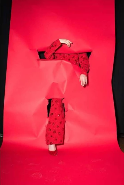 Person in patterned red jacket with red shoes emerging from torn red paper, set against black background, fashion concept.