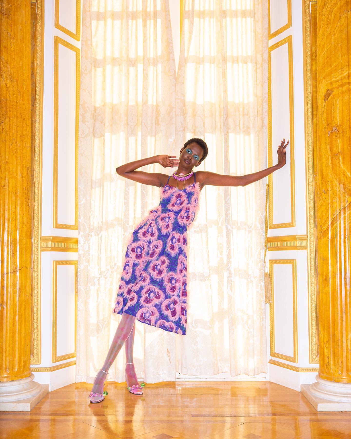 A tall, slender woman posing in a floral dress in an opulent indoor setting with gold columns and high ceilings with cream-colored curtains.