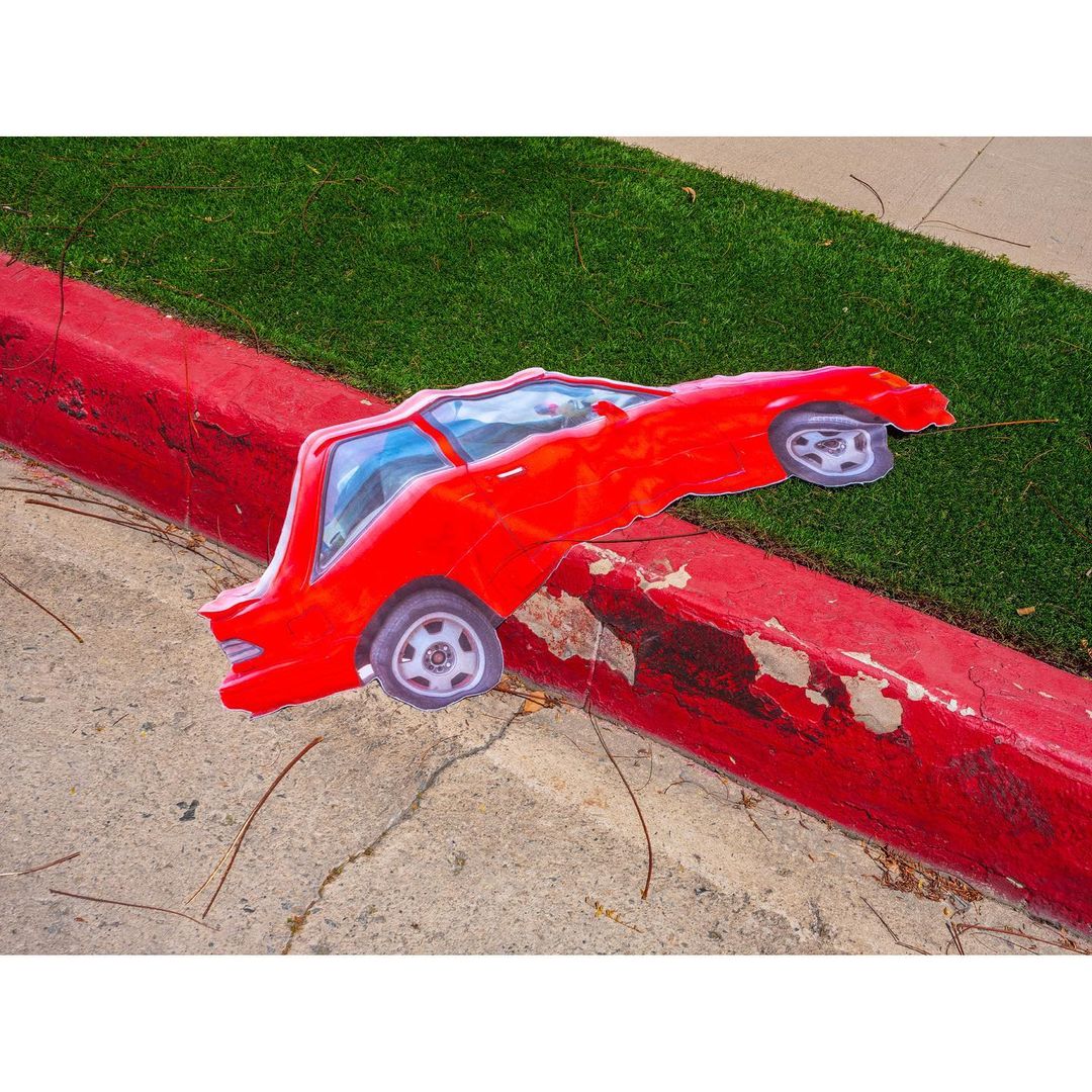 Red car artwork placed on concrete curb with grass background; vivid, playful automotive art.