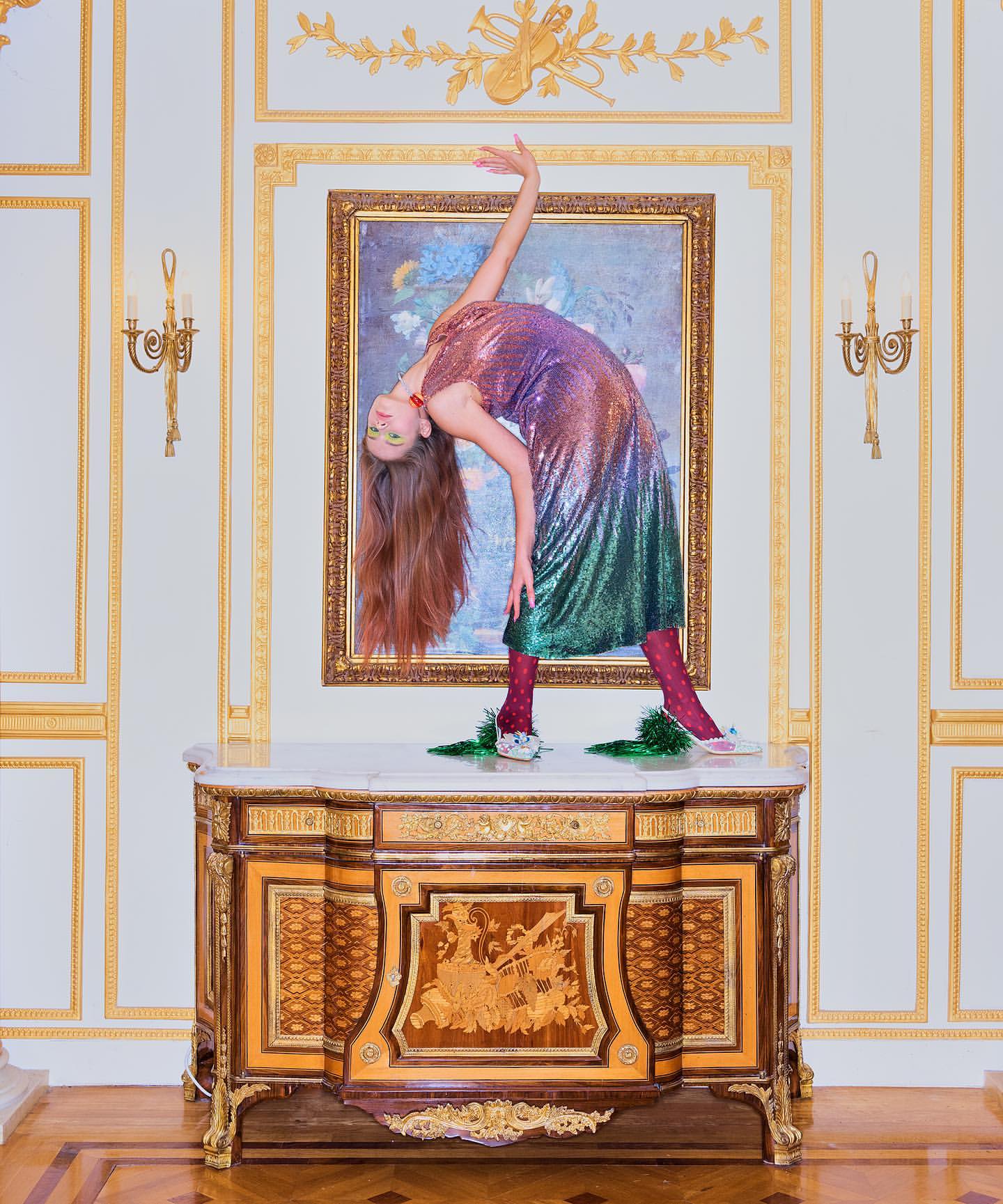 Young woman in colorful sequin dress on antique cabinet with ornate white and gold wall, performing a backbend in an interior setting.
