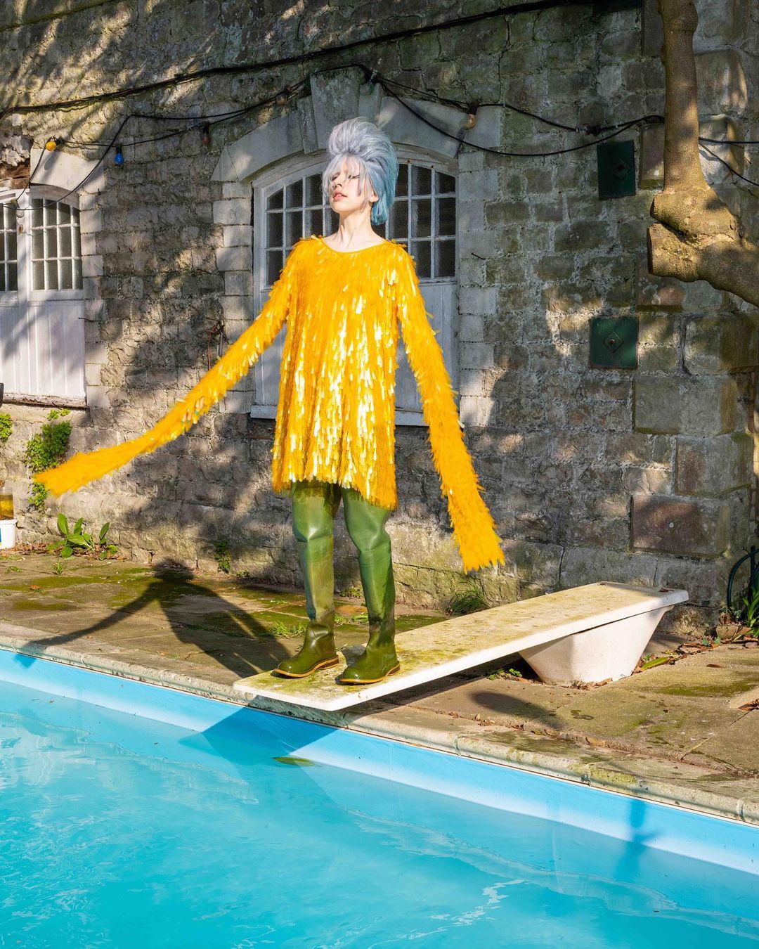 Model with elaborate grey hair and long-sleeved gold dress, standing on a diving board next to a pool, in front of a stone building.