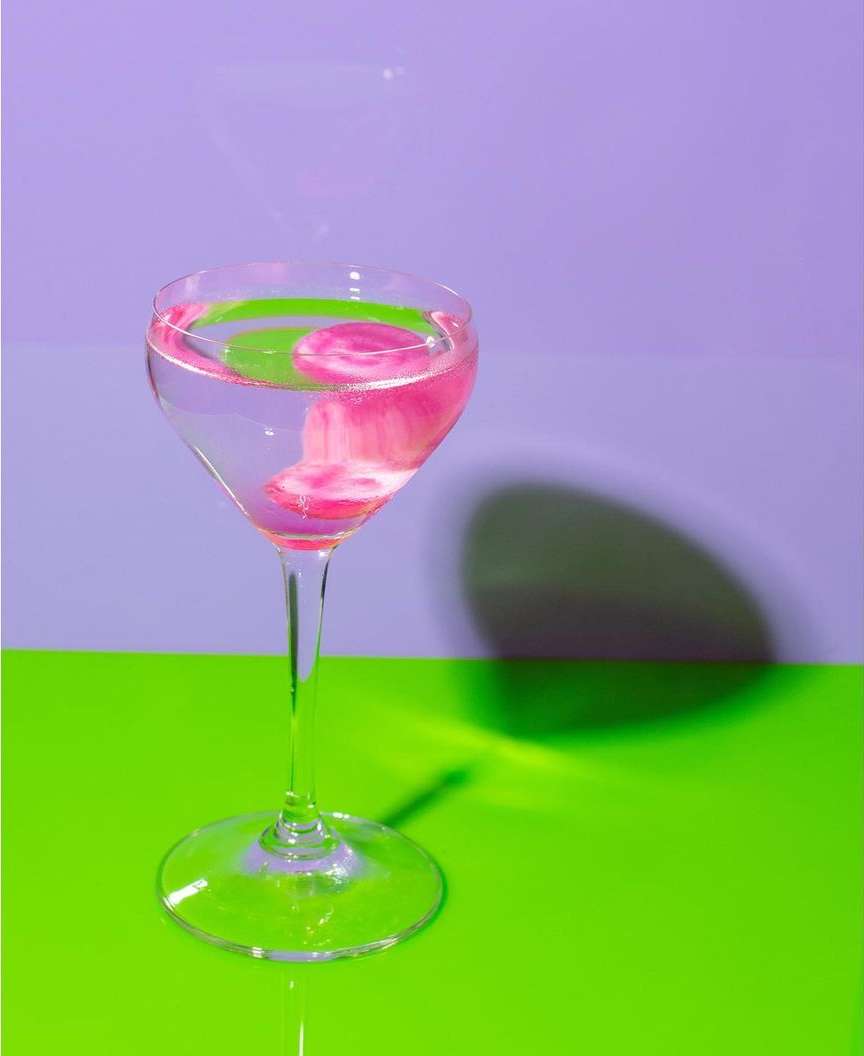 Elegant wine glass filled with vibrant pink liquid set against a contrasting green and purple background, casting a soft shadow.