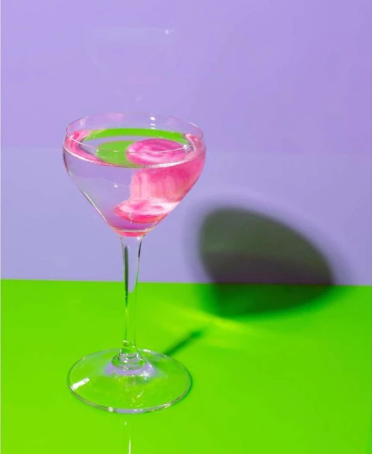 Elegant wine glass filled with vibrant pink liquid set against a contrasting green and purple background, casting a soft shadow.