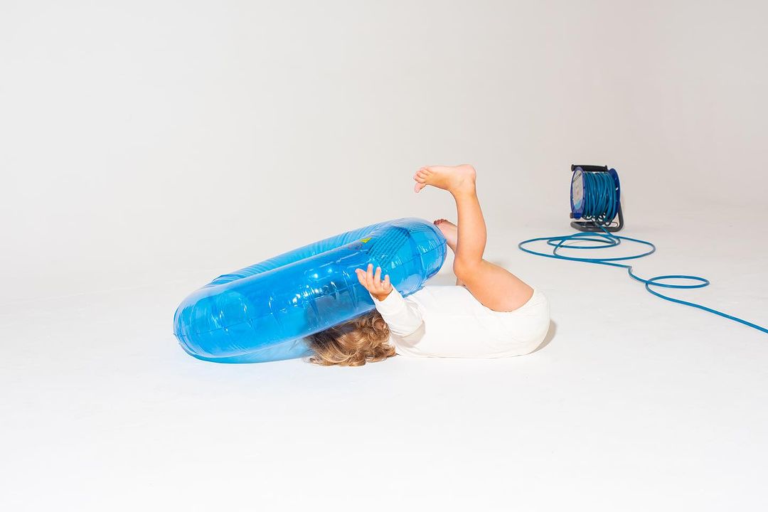 Child lies on a white studio floor with legs up and head under a large, blue inflatable inner tube; the background is simple and bright.