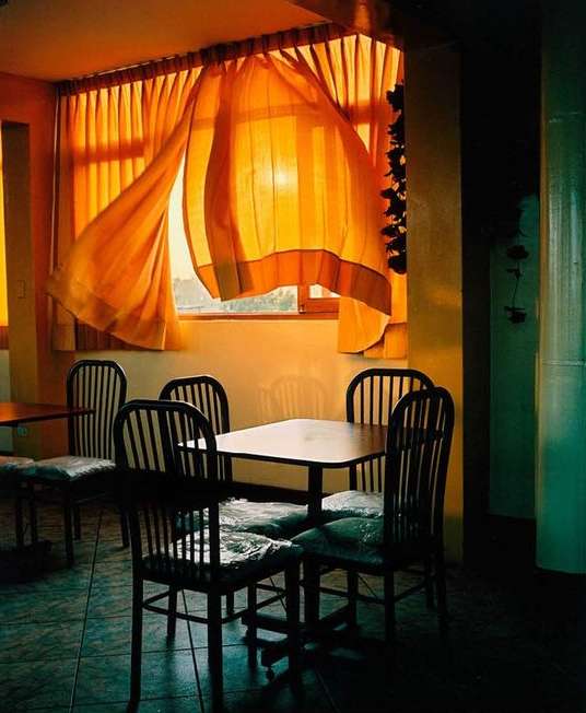 Sunlight streams through billowing curtains in an empty dining room, casting a warm glow on the tables and chairs.