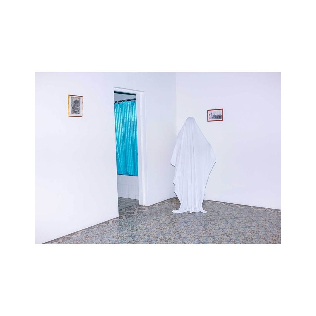A person covered in a white sheet stands in a room with patterned flooring and white walls, creating a ghostly presence.
