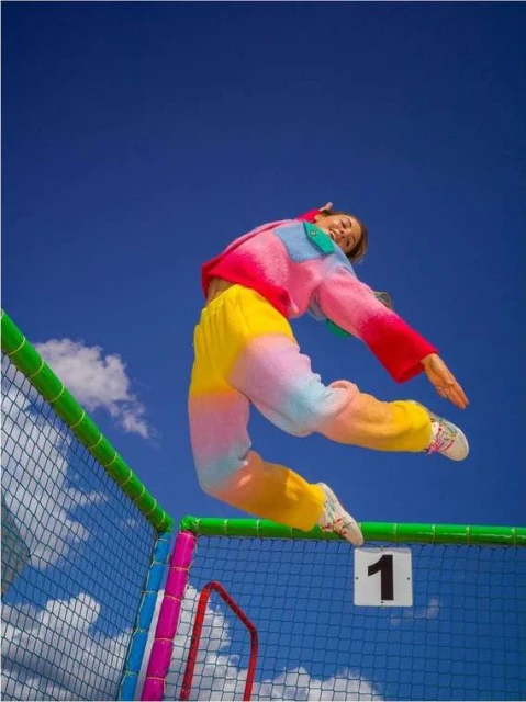 A young adult female leaps against a blue sky backdrop, wearing a vibrant pink and yellow sweater and tie-dye pants, action shot.