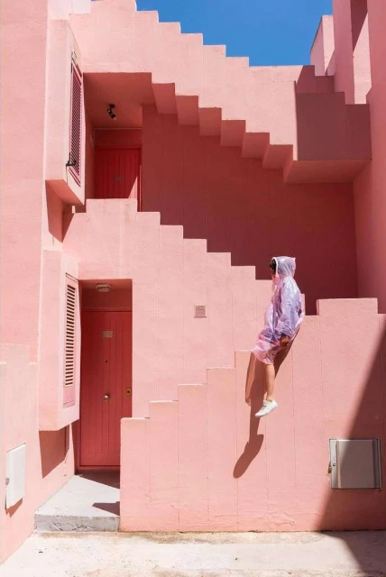 Woman in translucent hooded raincoat sitting on the steps of a pink building with minimalist architectural details on a sunny day.
